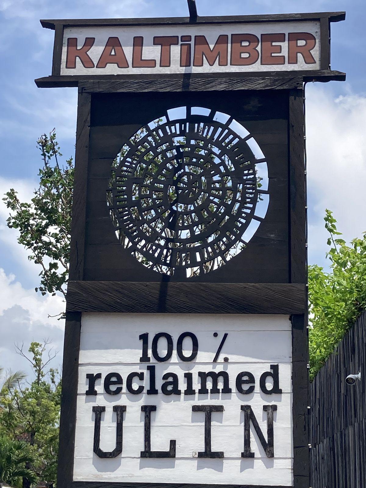 The roadside sign at Kaltimber near Ubud in Bali, reading Kaltimber, 100% reclaimed ulin, with a carved spiral of ironwood blocks